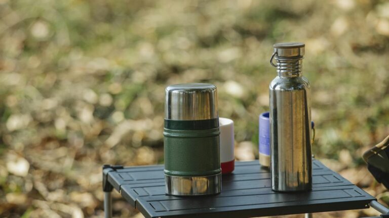 A tranquil outdoor camping scene featuring a portable table with a stainless steel thermos and cups amidst nature.