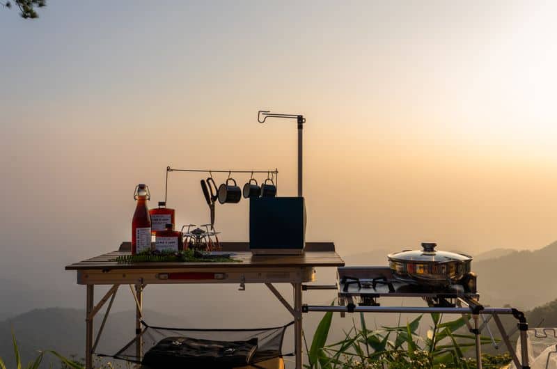 Campingurlaub planen: Unsere Tipps für Packliste, Ausrüstung & Kosten outdoor kitchen setup with pots and bottles overlooking a serene mountain range at sunset. 15306971 Faltbare Campingküche kaufen: Welche ist wirklich praktisch und robust?